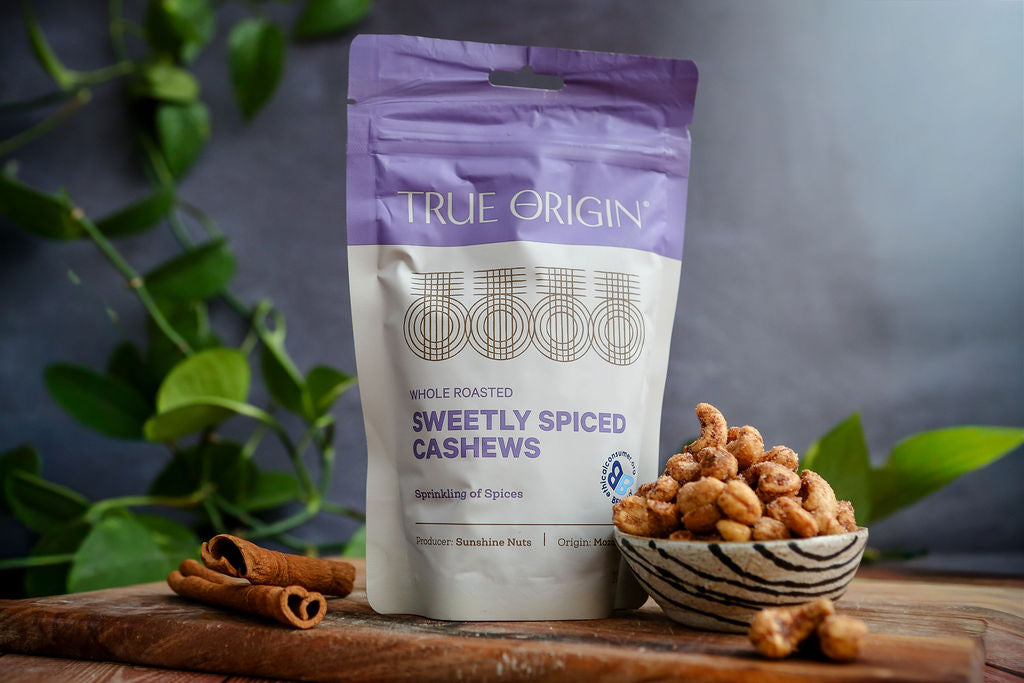 True Origin sweetly spiced cashews packaging with cashews on a wooden board, with a bowl of cashews and cinnamon in the background.