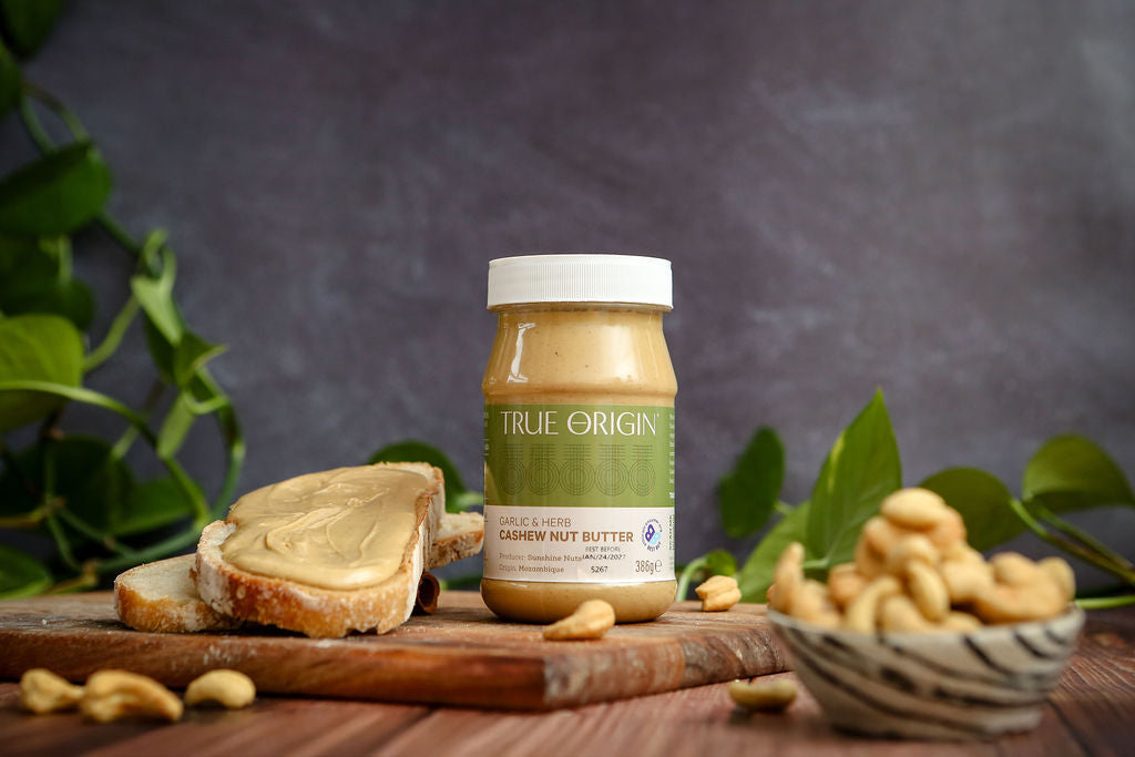 Jar of True Origin cashew butter with bread and cashews on a wooden surface