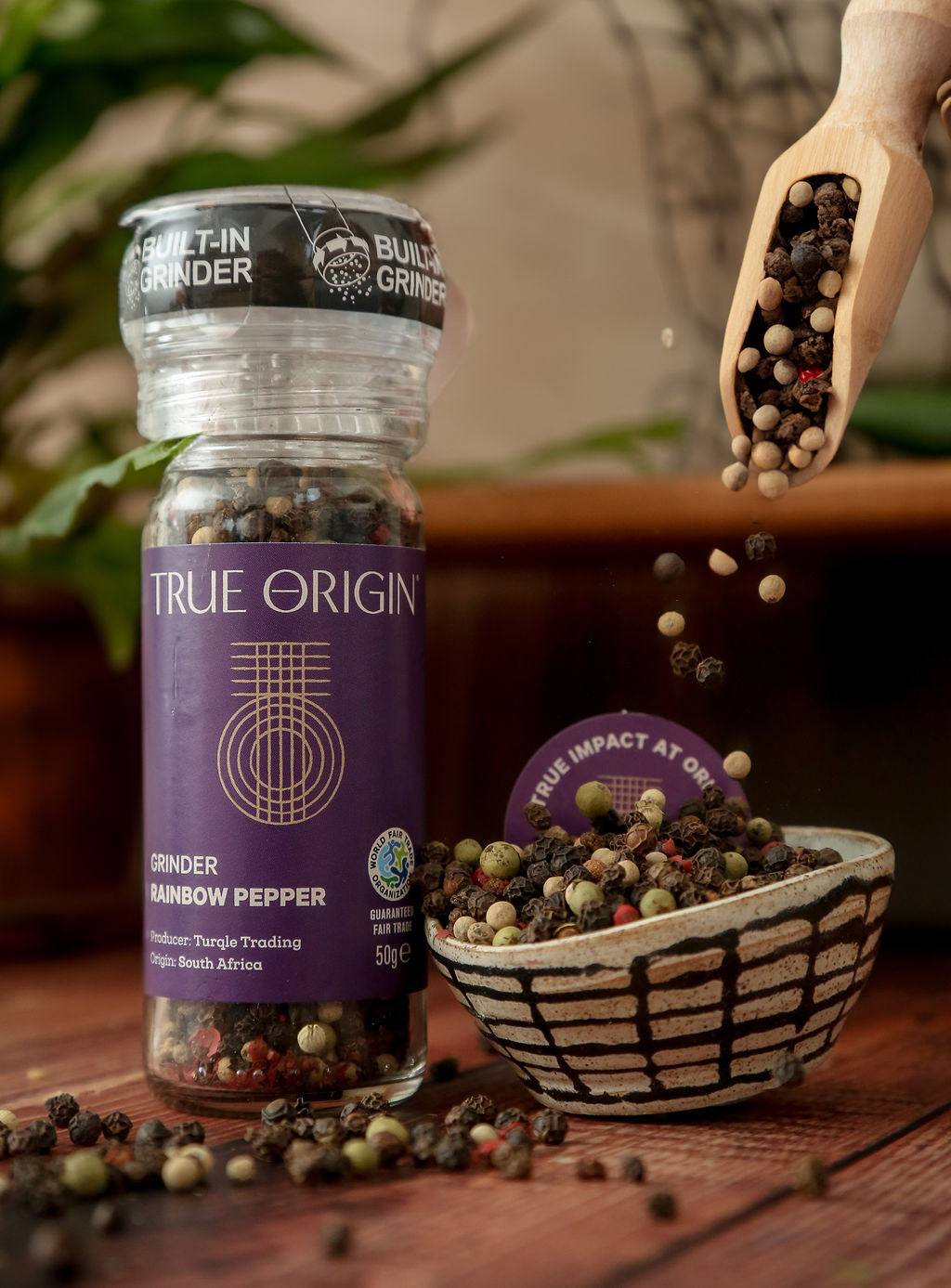 True Origin pepper grinder with a bowl of fair trade rainbow peppercorns on a wooden surface.
