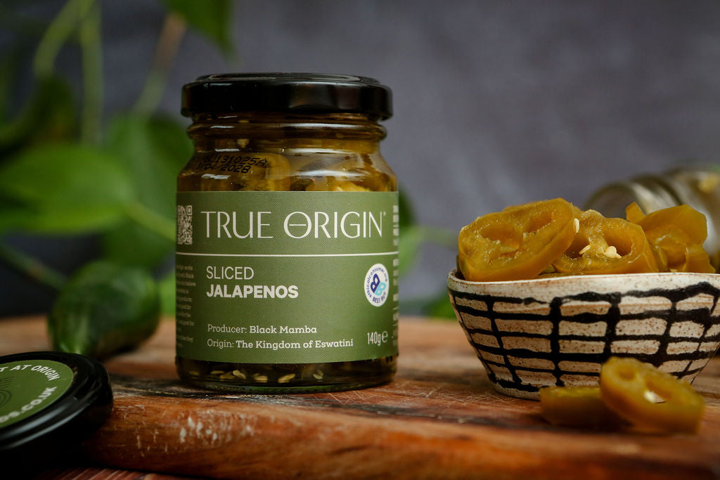 Jars of True Origin pickled jalapeños on a wooden board, along with a bowl of contents, with green leaves in the background.