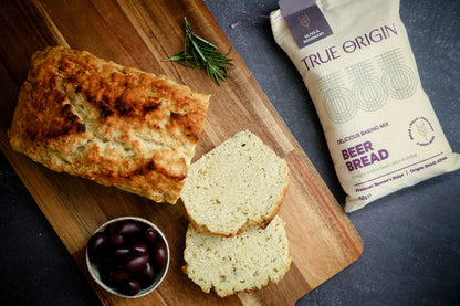 Loaf of Olive & Rosemary Beer Bread with slices on a wooden board next to a package of True Origin beer bread.