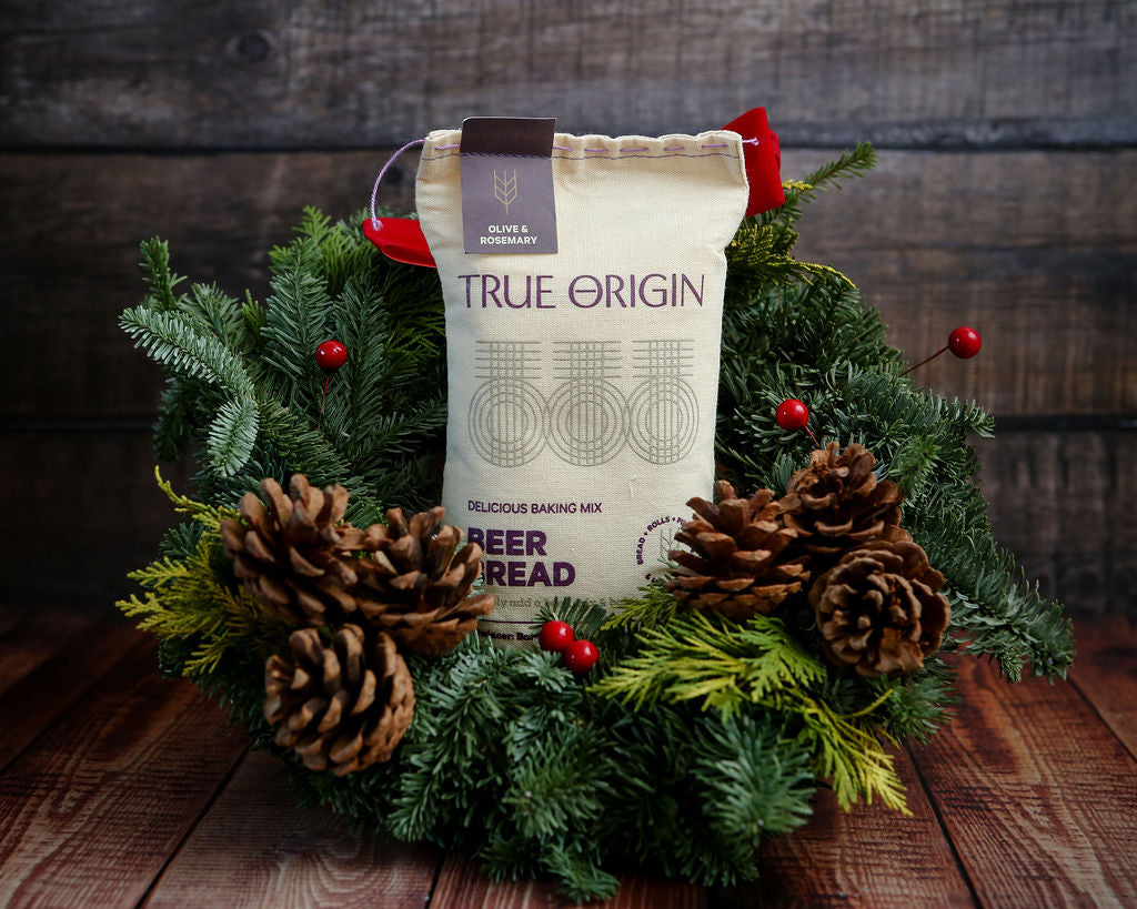 True Origin Olive & Rosemary Beer Bread mix package surrounded by festive greenery and pinecones on a wooden surface.
