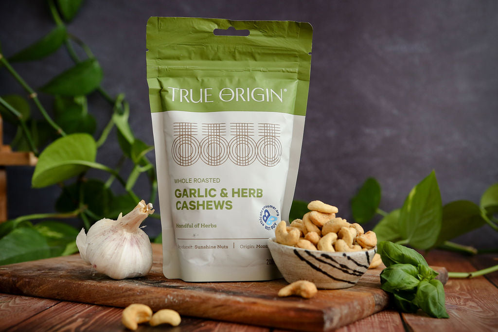 True Origin Garlic & Herb Cashews packaging on a wooden surface with cashews and garlic.