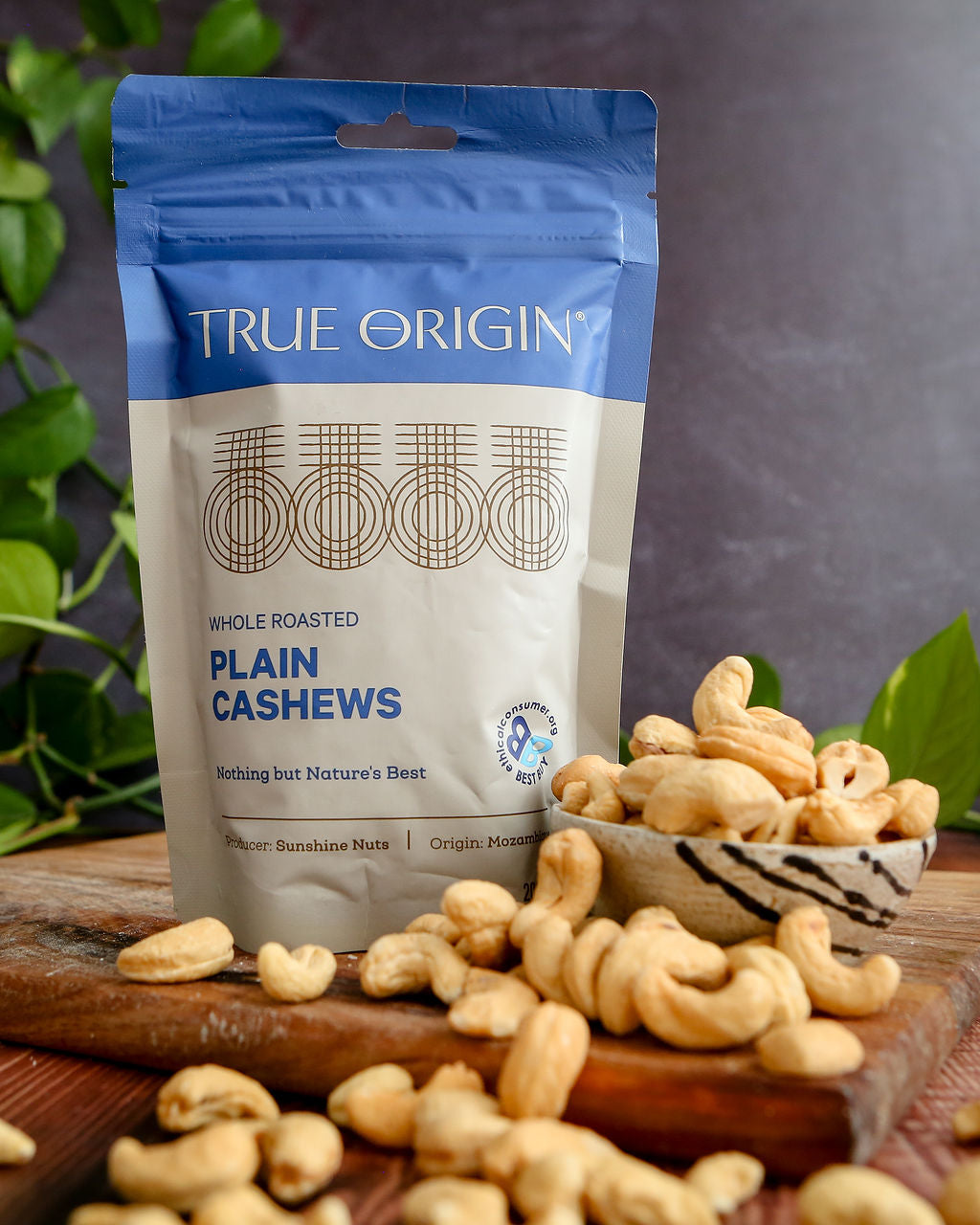 Bag of True Origin plain cashews on a wooden surface with cashews scattered around.