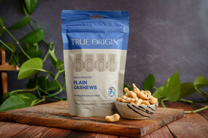 True Origin plain cashews packaging on a wooden board with cashews around, against a dark background with greenery.