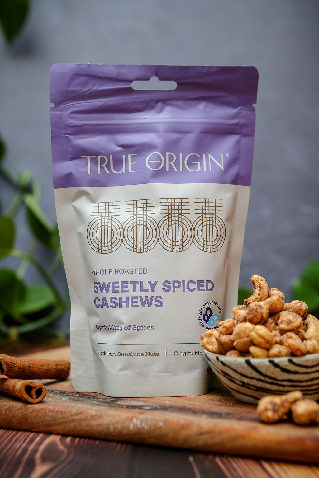 Bag of True Origin sweetly spiced cashews on a wooden surface with cashews and cinnamon sticks.
