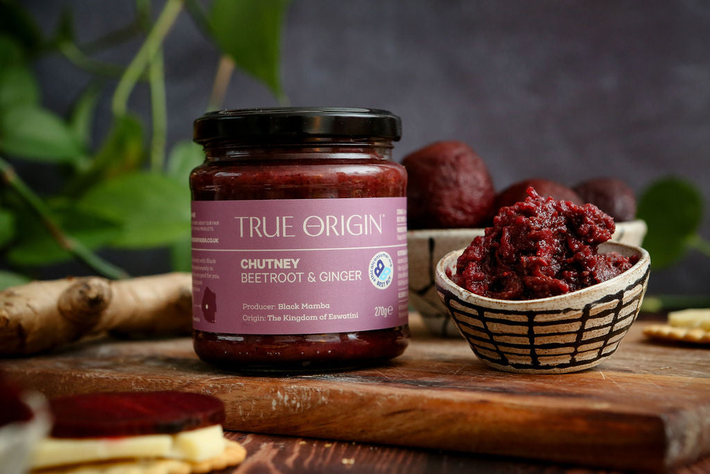 Jar of True Origin chutney with a small bowl of chutney on a wooden surface and beetroot, ginger and leaves in the background.