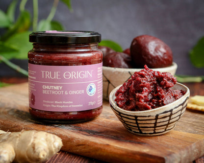 Jar of True Origin chutney with a small bowl of chutney on a wooden surface and beetroot, ginger and leaves in the background.