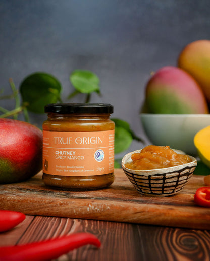 Jar of True Origin Chutney Spicy Mango on a wooden board with mangoes and chilies in the background.
