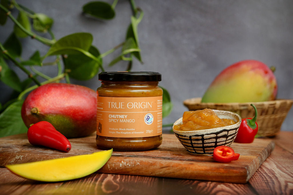 Jar of True Origin Chutney Spicy Mango on a wooden board with mangoes and chilies in the background.