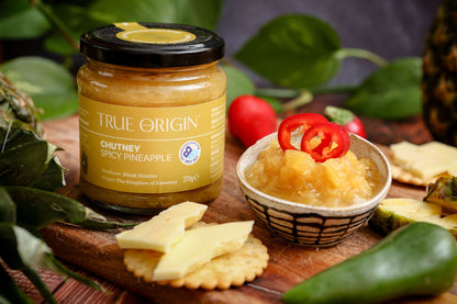 Jar of True Origin Spicy Pineapple Chutney on a wooden board with crackers, cheese and product.