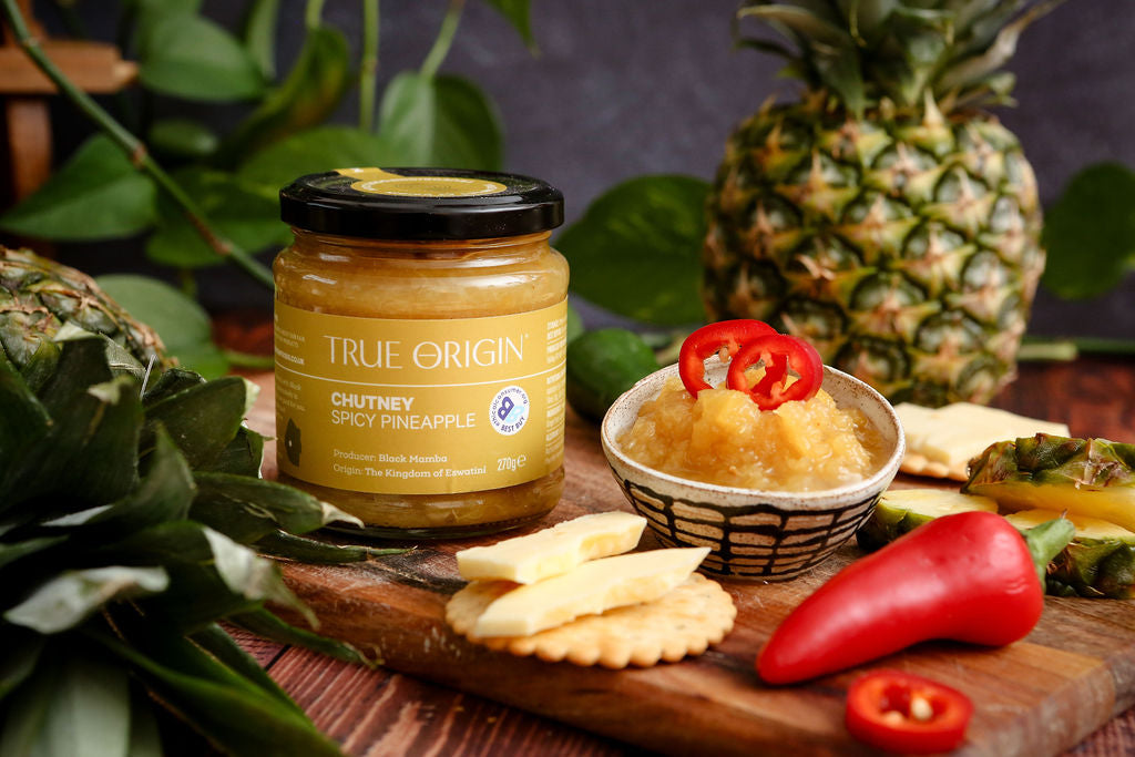 Jar of True Origin Spicy Pineapple Chutney on a wooden board with crackers, cheese and product.