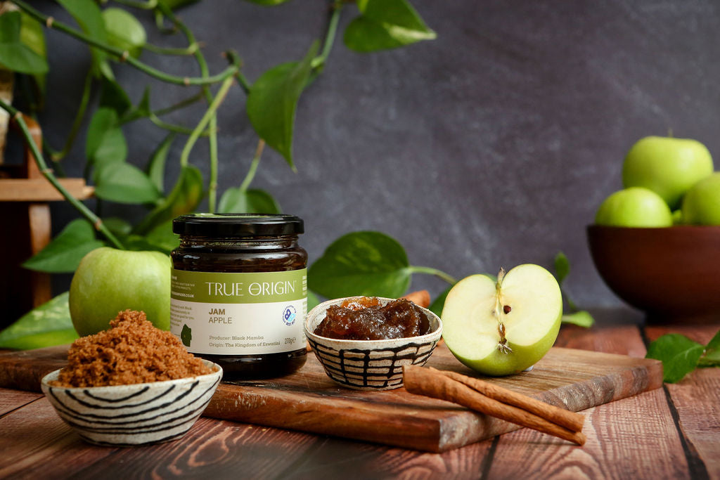 Jar of 'True Origin' jam on a wooden board with apples, cinnamon sticks and a bowl of product.