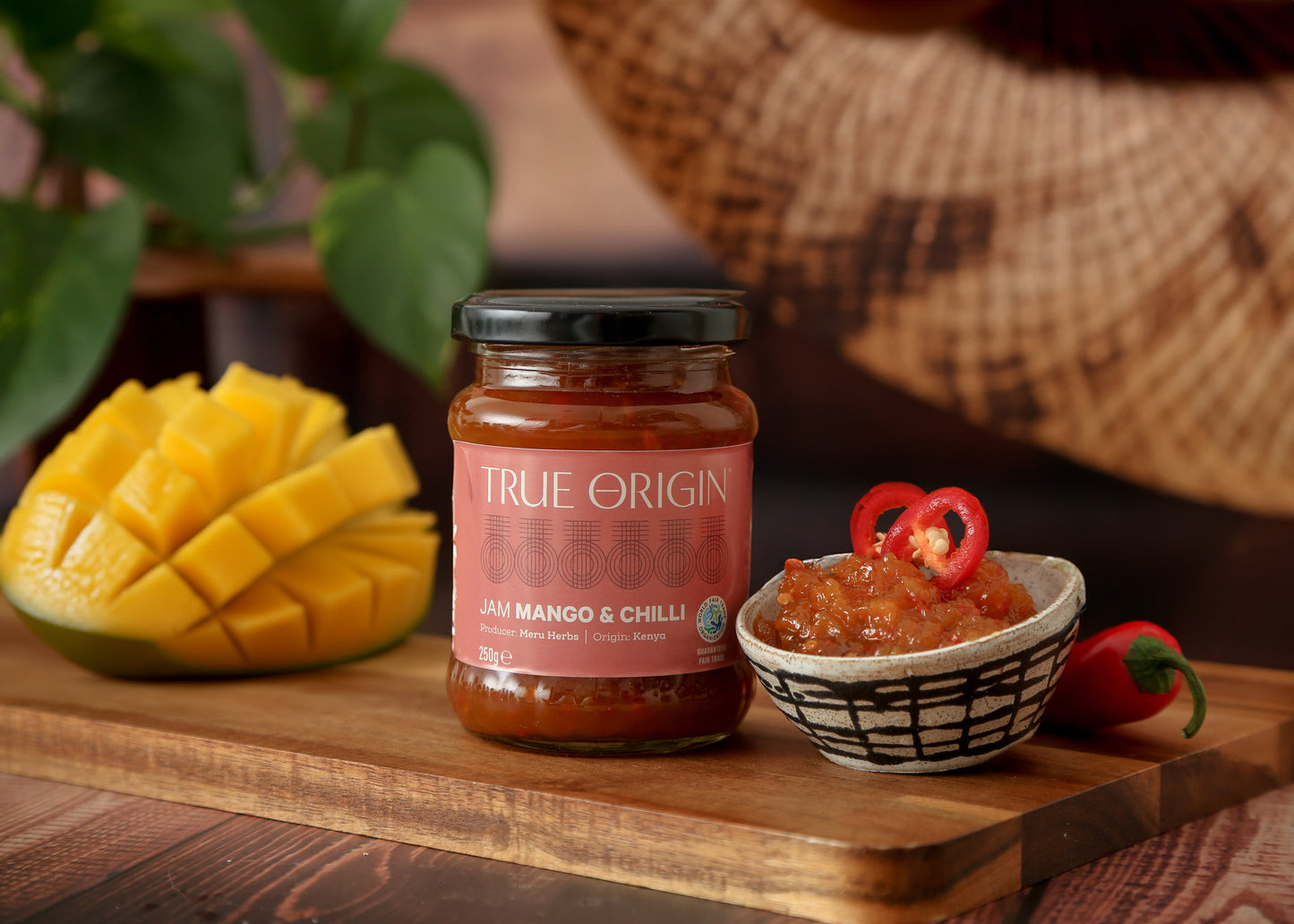 A jar of Mango & Chilli Jam with the label 'TRUE ORIGIN' displayed on it.  It is sitting next to a bowl filled with mango and chilli jam, some fresh chillies and a bowl of mangoes.