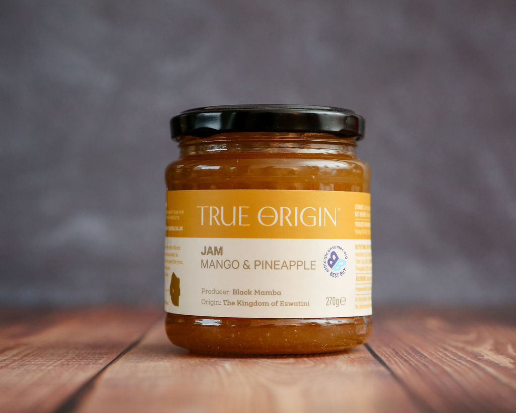 Jar of True Origin mango and pineapple jam on a wooden surface with a gray background