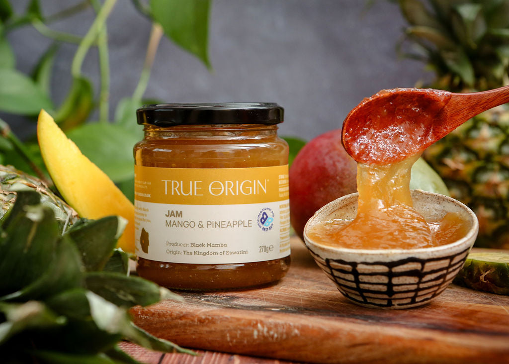 Jar of True Origin Mango & Pineapple jam with a spoonful of jam on a wooden board.