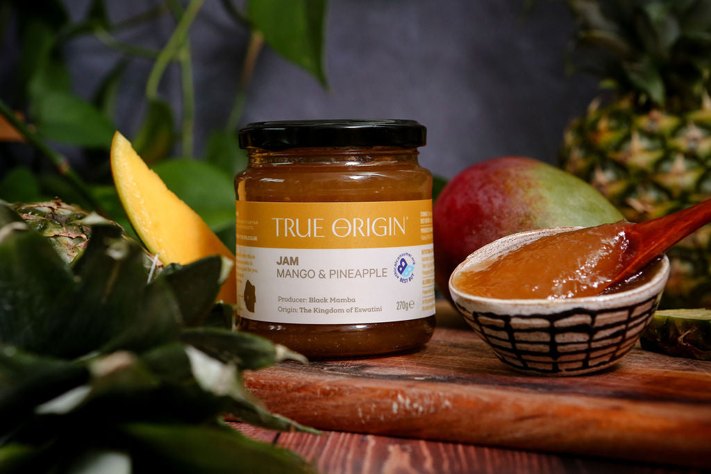 Jar of True Origin Mango & Pineapple Jam on a wooden board with fruits in the background