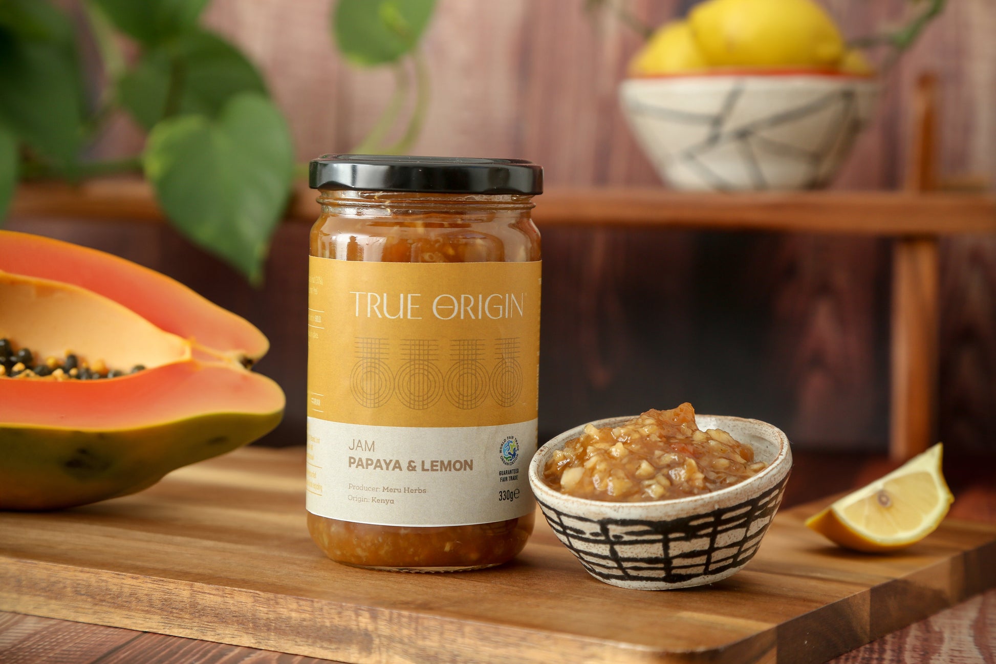 A glass jar of Papaya & Lemon Jam with a label that reads 'TRUE ORIGIN' and 'PAPAYA & LEMON JAM'. The jar is sealed with a black lid.  It is sitting on a wooden chopping board next to a bowl filled with papaya and lemon jam and a cut open fresh papaya and a bowl of lemons in the background.