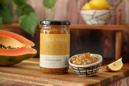 A glass jar of Papaya & Lemon Jam with a label that reads 'TRUE ORIGIN' and 'PAPAYA & LEMON JAM'. The jar is sealed with a black lid.  It is sitting on a wooden chopping board next to a bowl filled with papaya and lemon jam and a cut open fresh papaya and a bowl of lemons in the background.