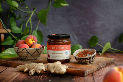 Jar of True Origin peach and ginger jam on a wooden surface with peaches and ginger root.