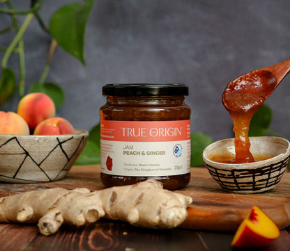 Jar of True Origin peach and ginger jam with a wooden spoon, peaches, and ginger on a wooden surface.