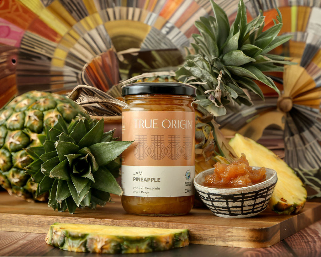 A jar of True Origin Pineapple Jam with an orange and white label next to a fresh pineapple and a bowl of pineapple jam.