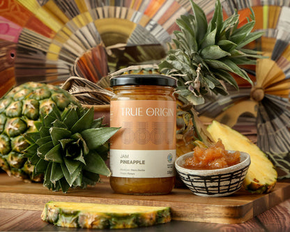 A jar of True Origin Pineapple Jam with an orange and white label next to a fresh pineapple and a bowl of pineapple jam.