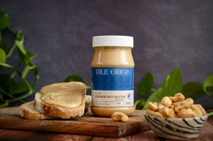Jar of True Origin cashew nut butter on a wooden board with bread and nuts, against a dark background.