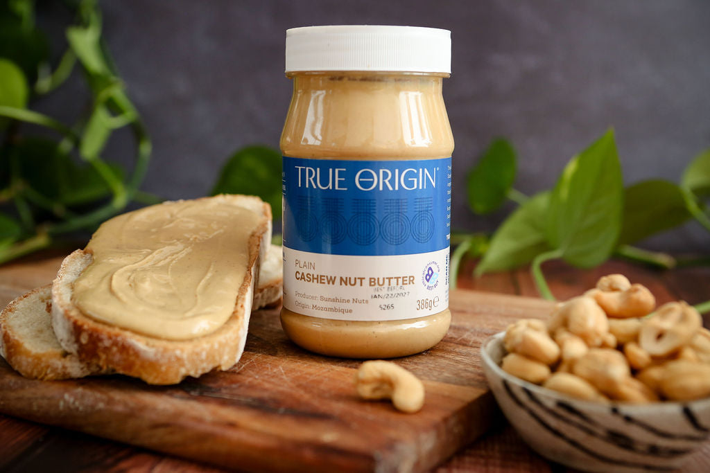Jar of True Origin cashew nut butter with spreaded toast and cashews on a wooden board.