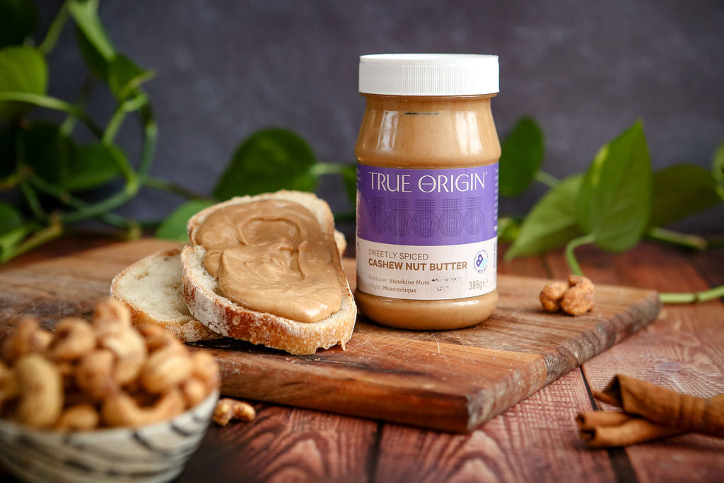 Jar of True Origin cashew nut butter on a wooden board with bread and nuts.