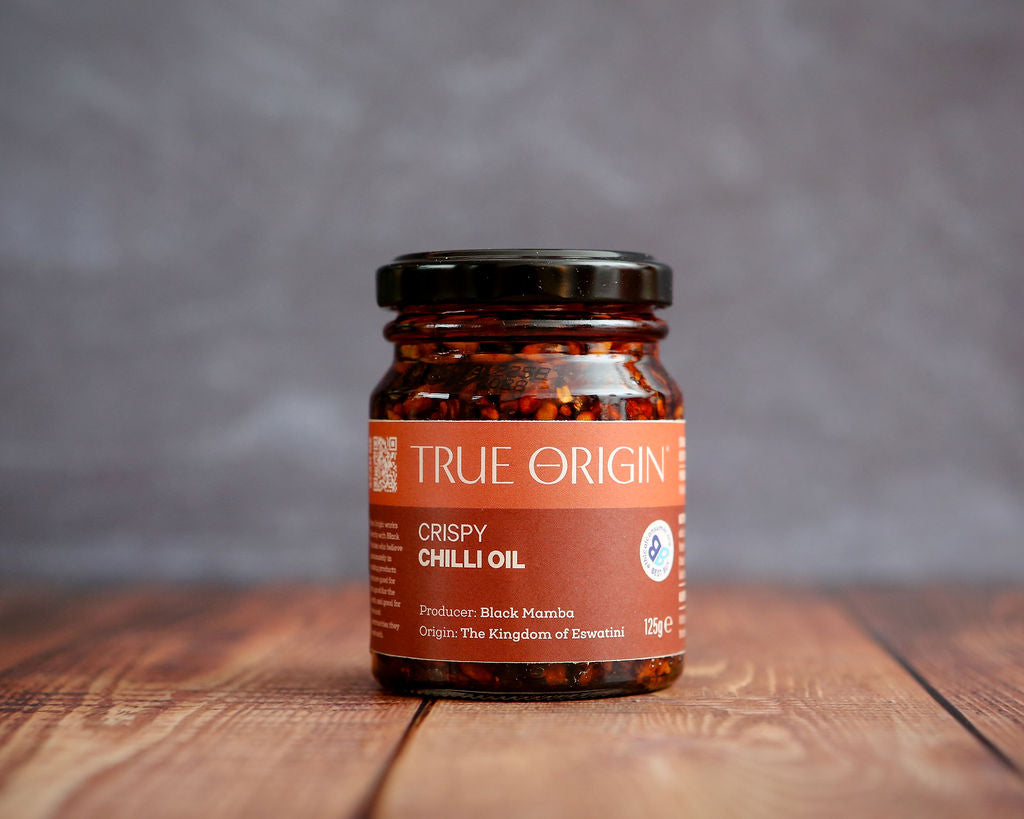 Jar of True Origin Crispy Chilli Oil on a wooden surface with a gray background