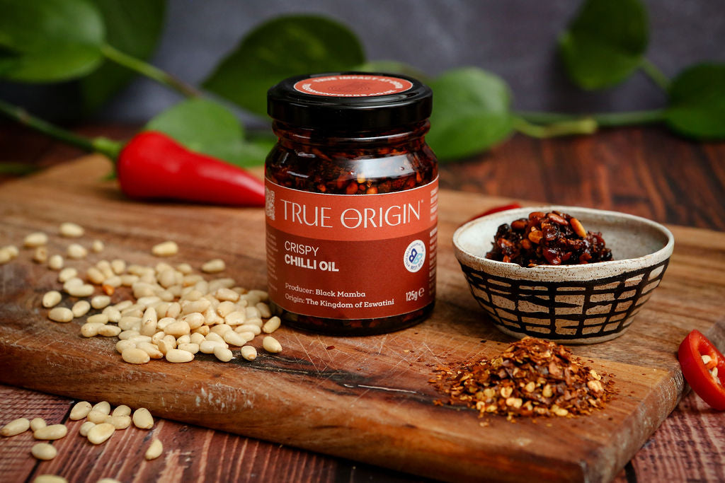 Jar of True Origin Crispy Chilli Oil with a bowl of contents sitting on a wooden chopping board.