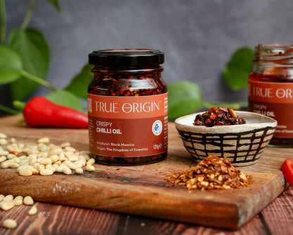 Jar of True Origin Crispy Chilli Oil on a wooden board with ingredients and a blurred background
