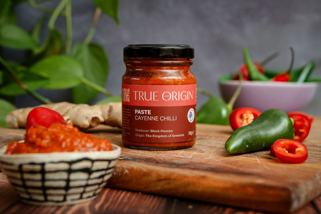 Jar of True Origins Paste Cayenne Chilli on a wooden board with peppers and product.