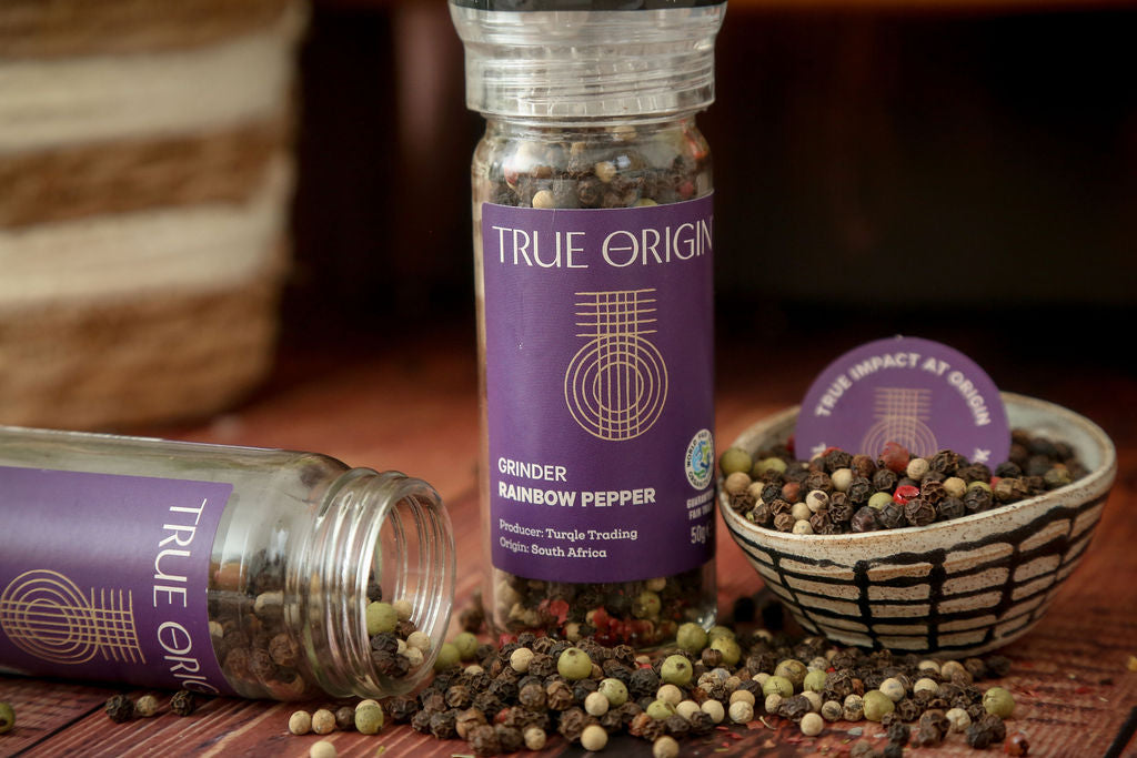 True Origin Rainbow Pepper grinder with open lid beside a bowl of rainbow peppercorns on a wooden surface