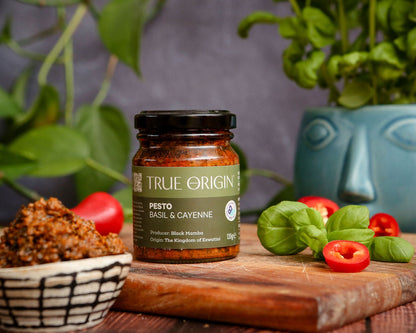 Jar of True Origin basil and cayenne pesto on a wooden cutting board with fresh basil and chillies and a bowl of contents.
