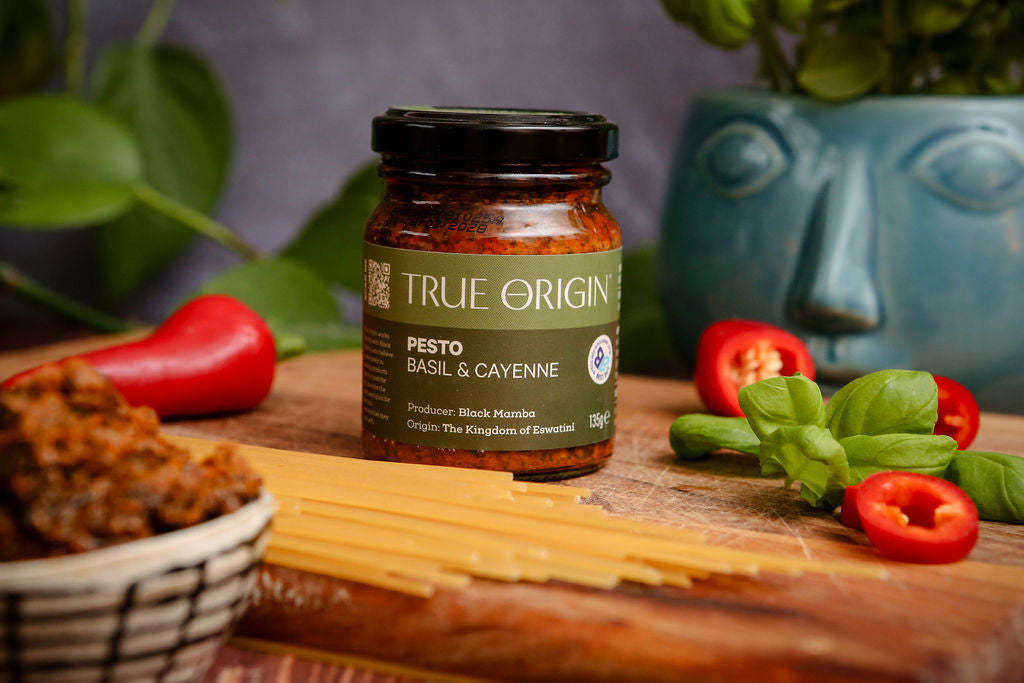 Jar of True Origin basil and cayenne pesto on a wooden cutting board with fresh basil and chillies and a bowl of contents.