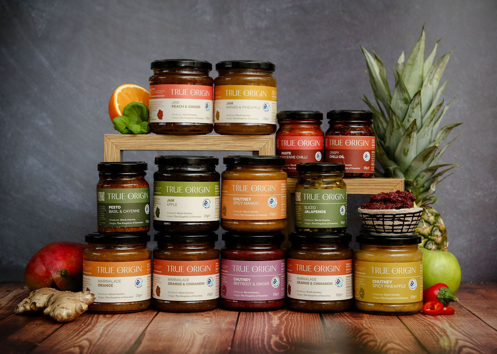 Assorted jars of True Origin products, by Black Mamba, on a wooden surface with fruits and vegetables.