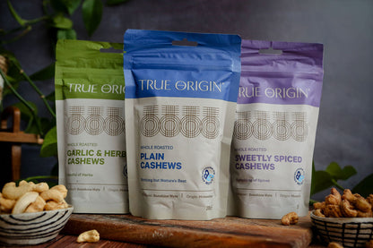 Three packages of True Origin cashews on a wooden board with nuts around, against a blurred natural background.