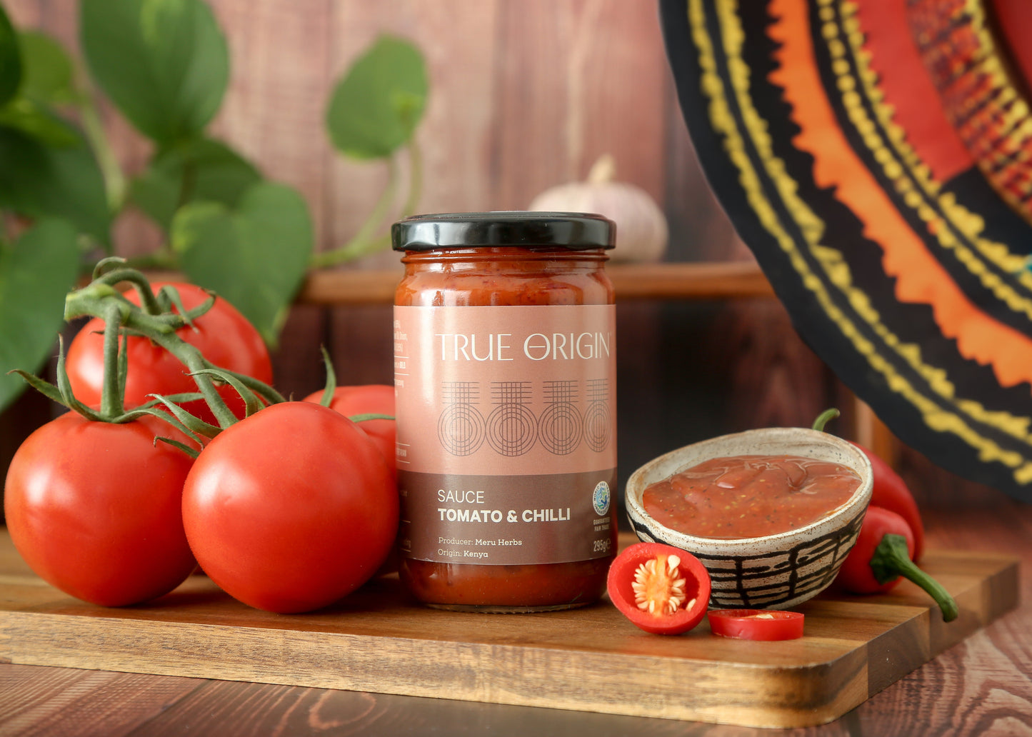 A jar of True Origin brand Tomato & Chilli sauce produced by Meru Herbs in Kenya.  The jar is sitting on a wooden chopping board next to some fresh tomatoes and fresh chillies and a bowl containing that tomato and chilli sauce.