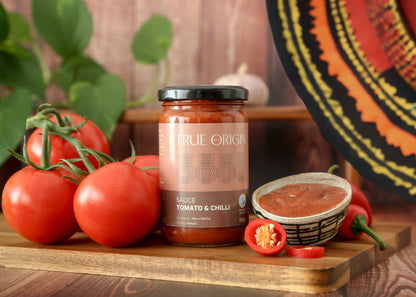 A jar of True Origin brand Tomato & Chilli sauce produced by Meru Herbs in Kenya.  The jar is sitting on a wooden chopping board next to some fresh tomatoes and fresh chillies and a bowl containing that tomato and chilli sauce.