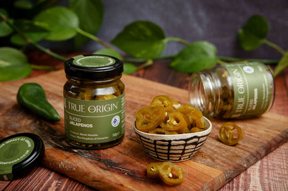 Jars of True Origin pickled jalapeños on a wooden board, along with a bowl of contents, with green leaves in the background.